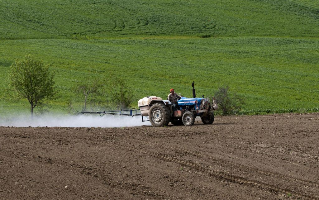 A tractor in a rural agricultural field conventionally spraying pesticides over the soil.